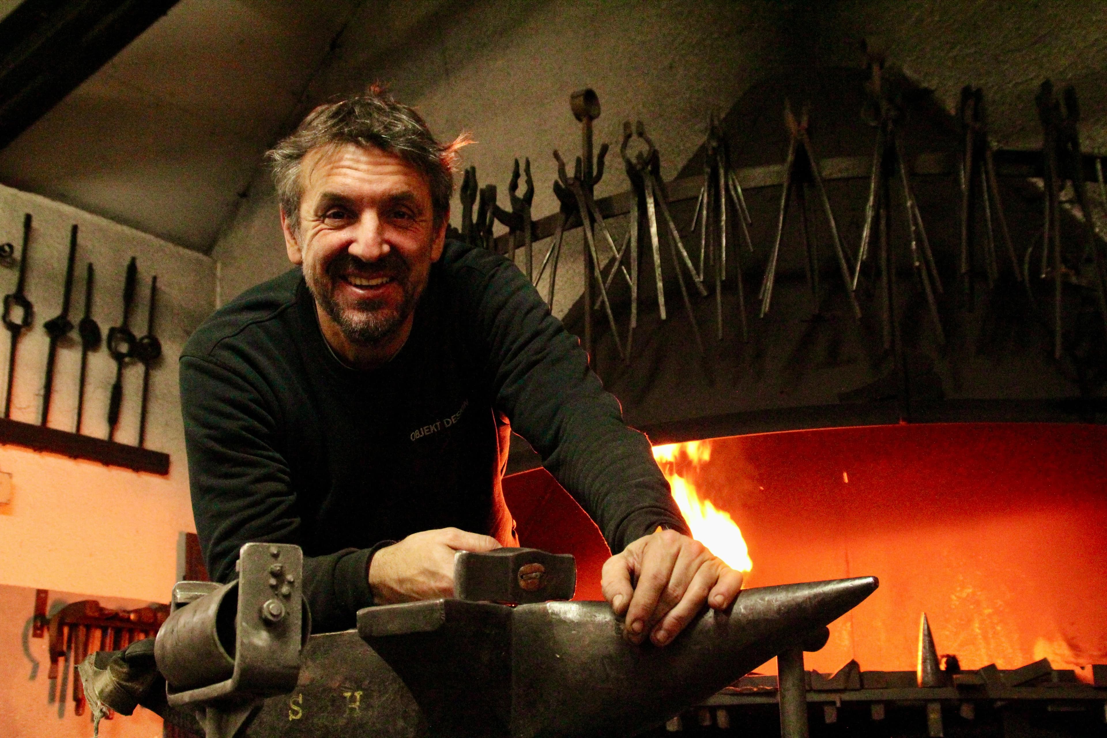 Thomas Bischoff, lachend über den Amboss gelehnt, dahinter das offene Feuer der Esse und eine Wand voller Zangen